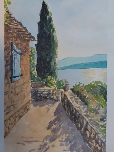 Ruelle de village méditerranéen avec un cyprès, un mur de pierre et le lac en contrebas.