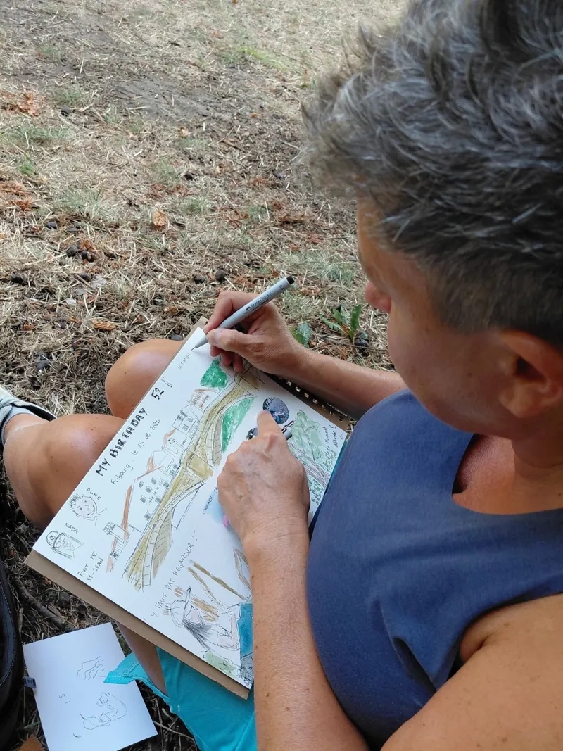 Une participante dessine à l'encre sur sa page de carnet, assise dans l'herbe.